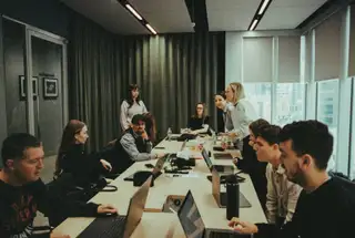A large group of coworkers sit around a conference table with laptops during a meeting, while two people stand and speak at the far end of the room.