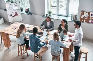 A group of six colleagues sit around a wooden table in a bright office, collaborating on documents and laptops during a team meeting.