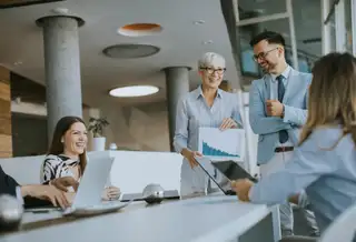 A group of colleagues in a modern office sit around a table smiling and discussing as an older woman standing at the front holds up a printed chart. A man beside her in a light blue suit is also smiling, and laptops are open in front of the seated team members.