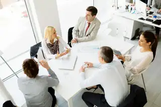 A group of five professionals sit around a white table in a bright modern office. They appear to be engaged in a meeting or discussion. Papers, a laptop, and glasses of water are on the table. Large windows let in natural light, and additional desks and computers are visible in the background.