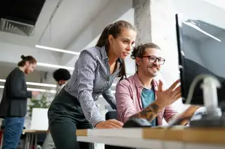 Two coworkers collaborate at a computer in a modern office, with a woman leaning in to look at the screen while a man gestures and smiles.