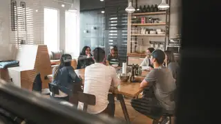 A group of coworkers sit around a wooden table in a modern office meeting room, talking and collaborating during a team discussion.